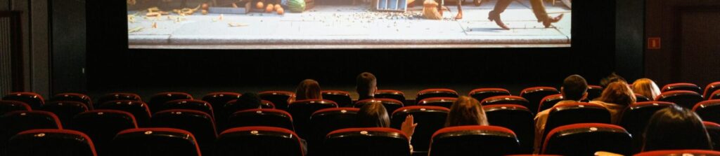 An audience sits in a dimly lit theater watching a vibrant animated film on a large cinema screen.