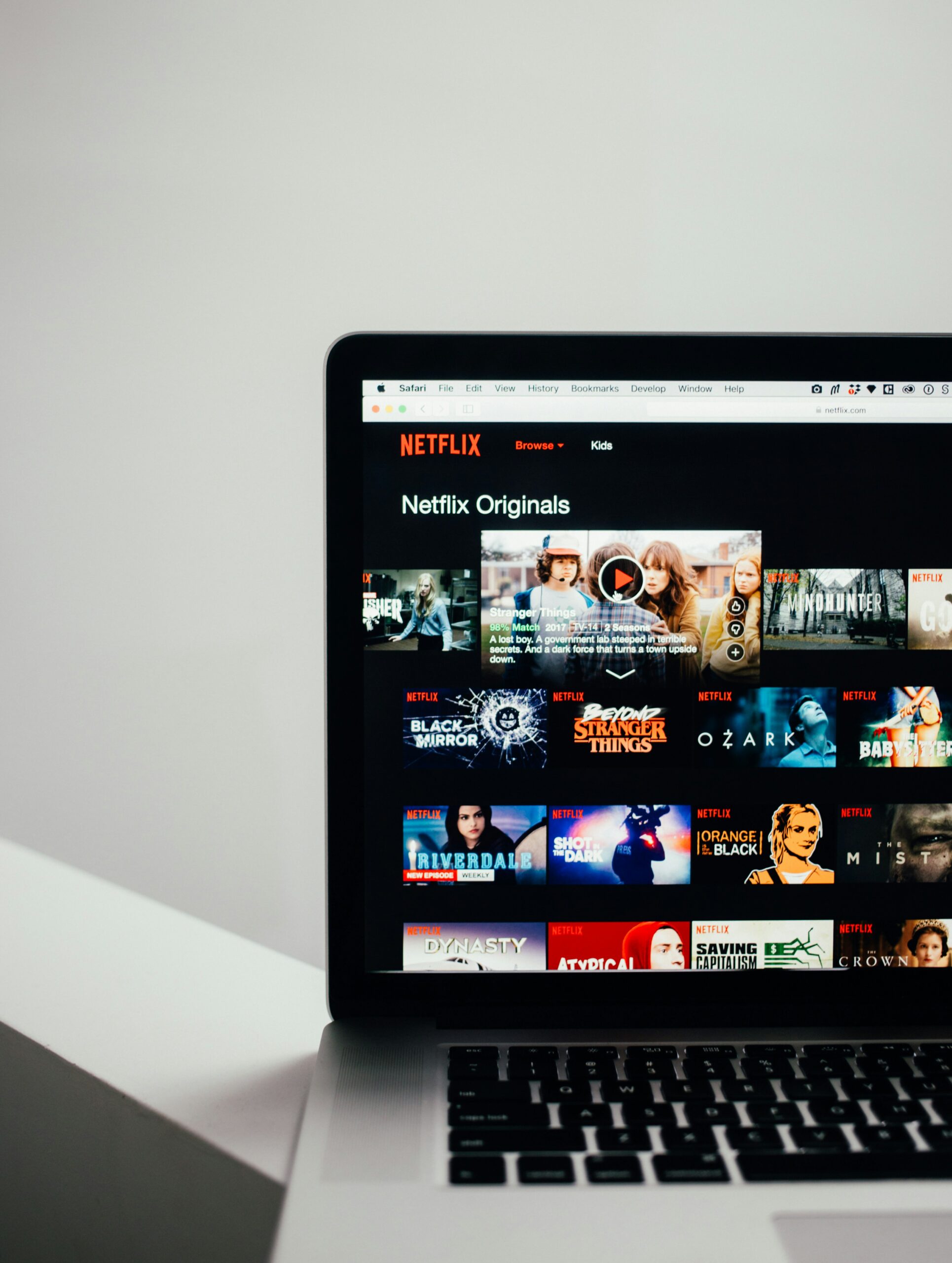 A MacBook laptop displays the Netflix Originals browsing page, showing a grid of show thumbnails including Stranger Things, Black Mirror, Ozark, Riverdale, and The Crown.