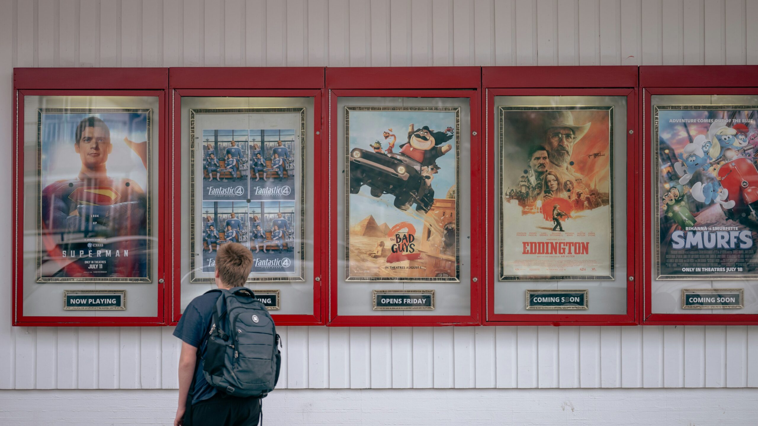 A young person with a backpack stands outside a movie theater, looking up at a row of film posters in red-framed display cases, including Superman, Fantastic Four, The Bad Guys 2, Eddington, and Smurfs.